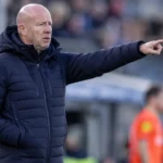 Henry van der Vegt in een donkere jas langs de zijlijn van een voetbalveld.