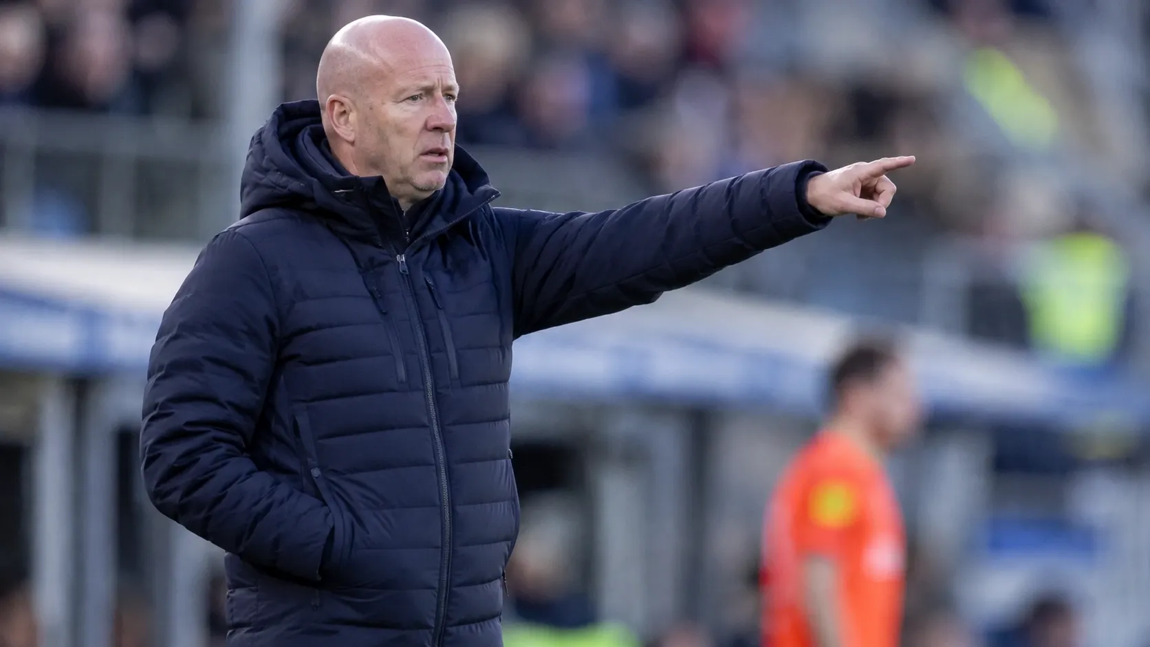 Henry van der Vegt in een donkere jas langs de zijlijn van een voetbalveld.