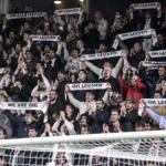 Een volle groep OH Leuven-supporters applaudisseert in de tribune met sjaals omhoog.