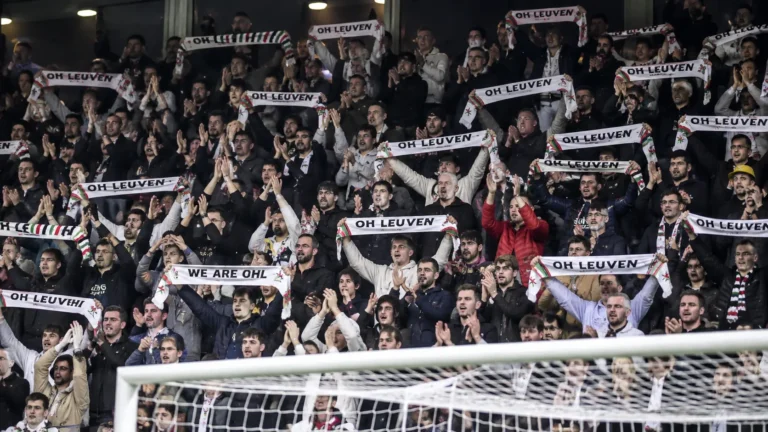 Een volle groep OH Leuven-supporters applaudisseert in de tribune met sjaals omhoog.