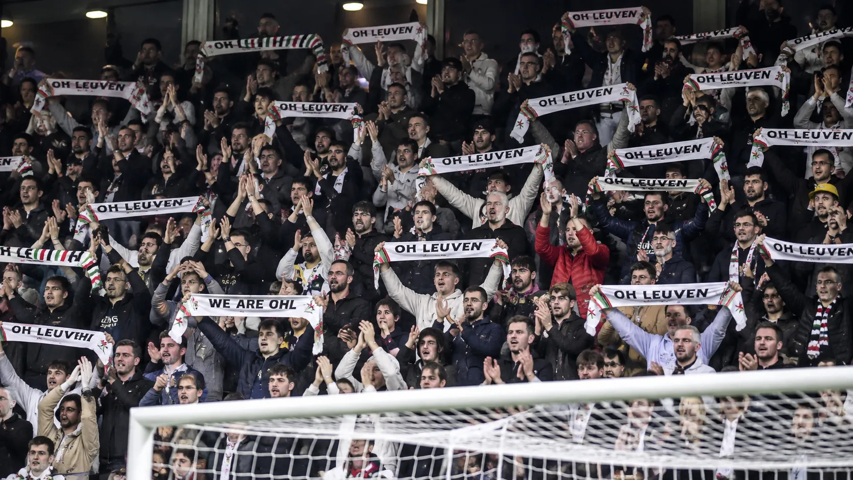 Een volle groep OH Leuven-supporters applaudisseert in de tribune met sjaals omhoog.
