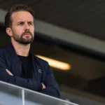 Een niet-spelende clubvertegenwoordiger in een donkere jas in een stadionomgeving.