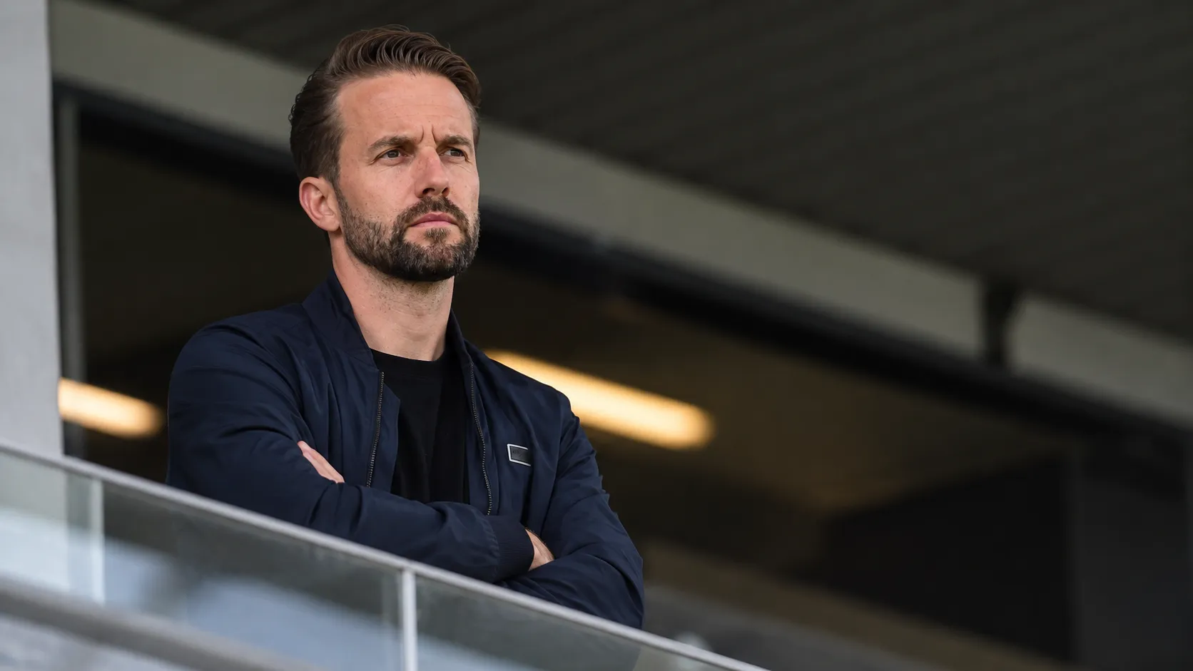 Een niet-spelende clubvertegenwoordiger in een donkere jas in een stadionomgeving.