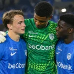 Drie spelers van KRC Genk steunen elkaar na een wedstrijd aan de rand van het veld.