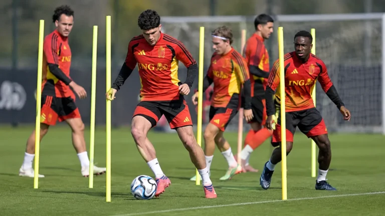 Een groep voetballers in rood-zwarte trainingskleding tijdens een oefening op het trainingsveld.
