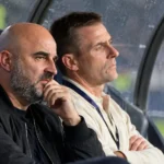 Twee niet-spelende clubmedewerkers kijken vanuit de dug-out aandachtig naar het veld.