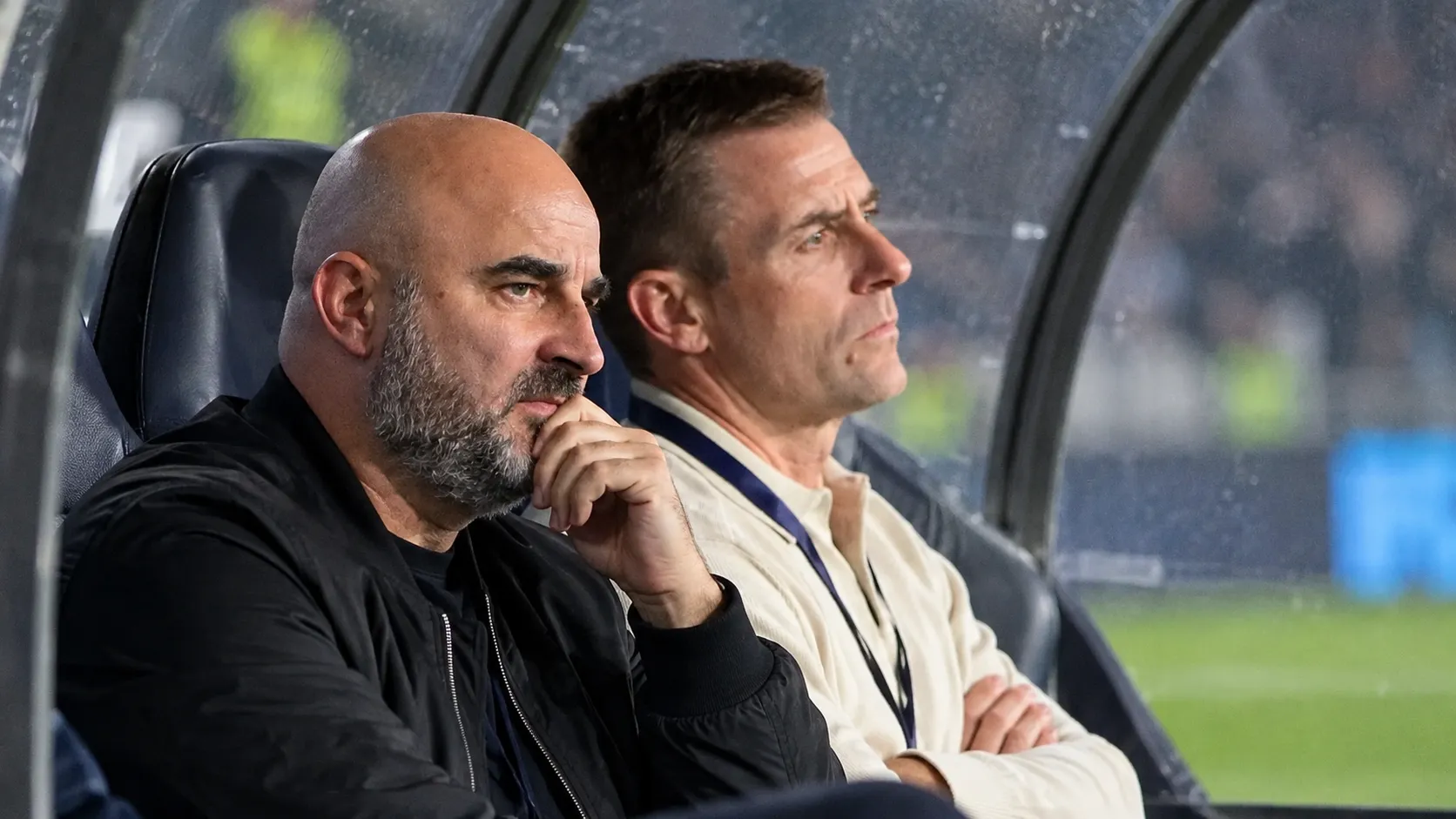 Twee niet-spelende clubmedewerkers kijken vanuit de dug-out aandachtig naar het veld.