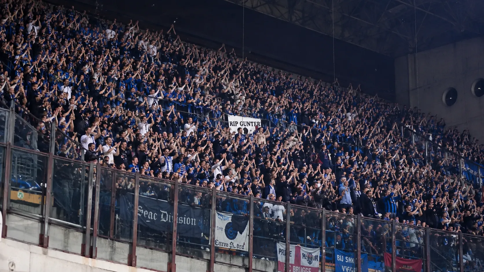 Een volle groep blauw-zwarte supporters applaudisseert in een stadiontribune.