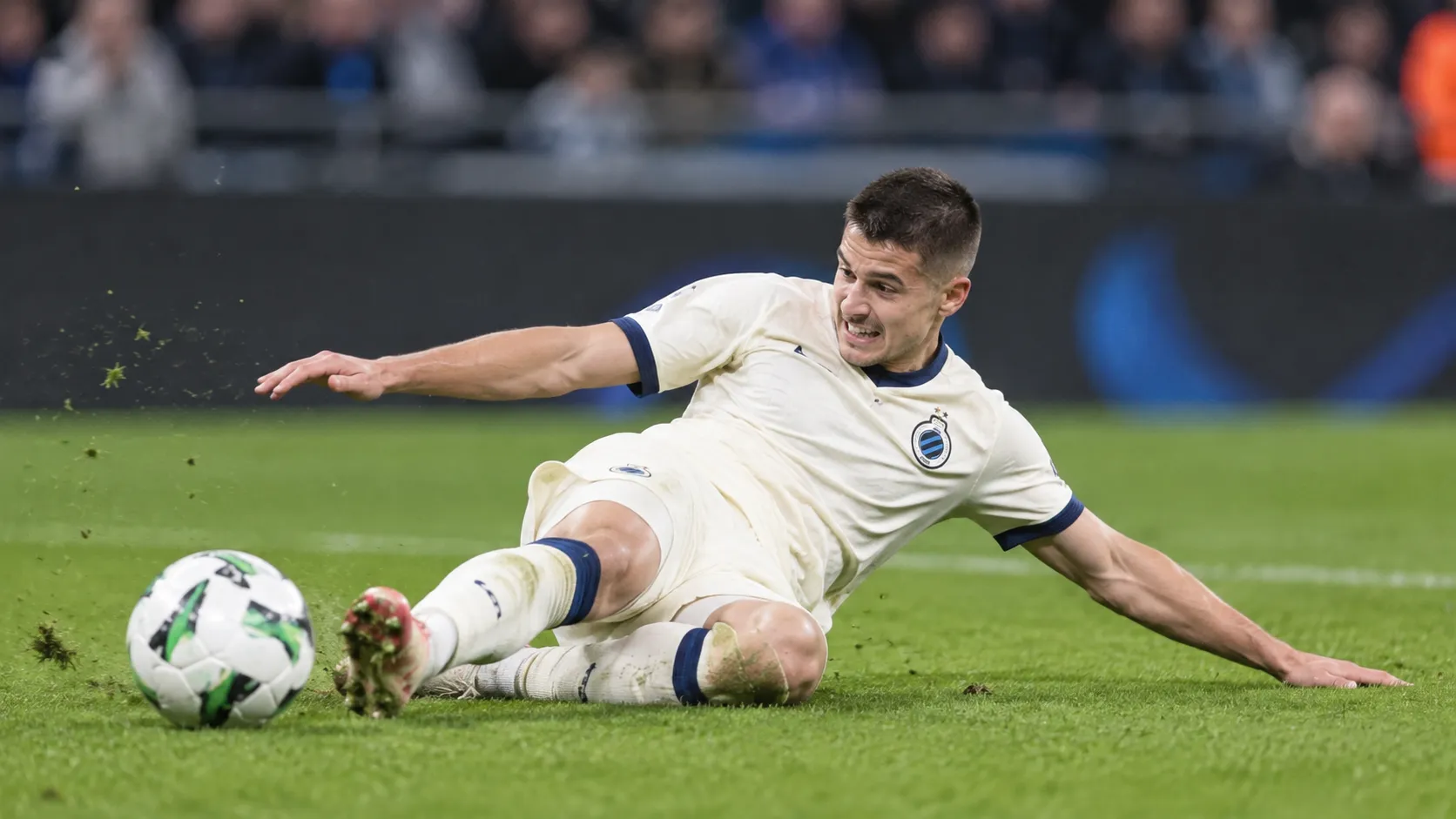 Een Club Brugge-speler in een licht wedstrijdtenue ligt op het gras tijdens een wedstrijd.