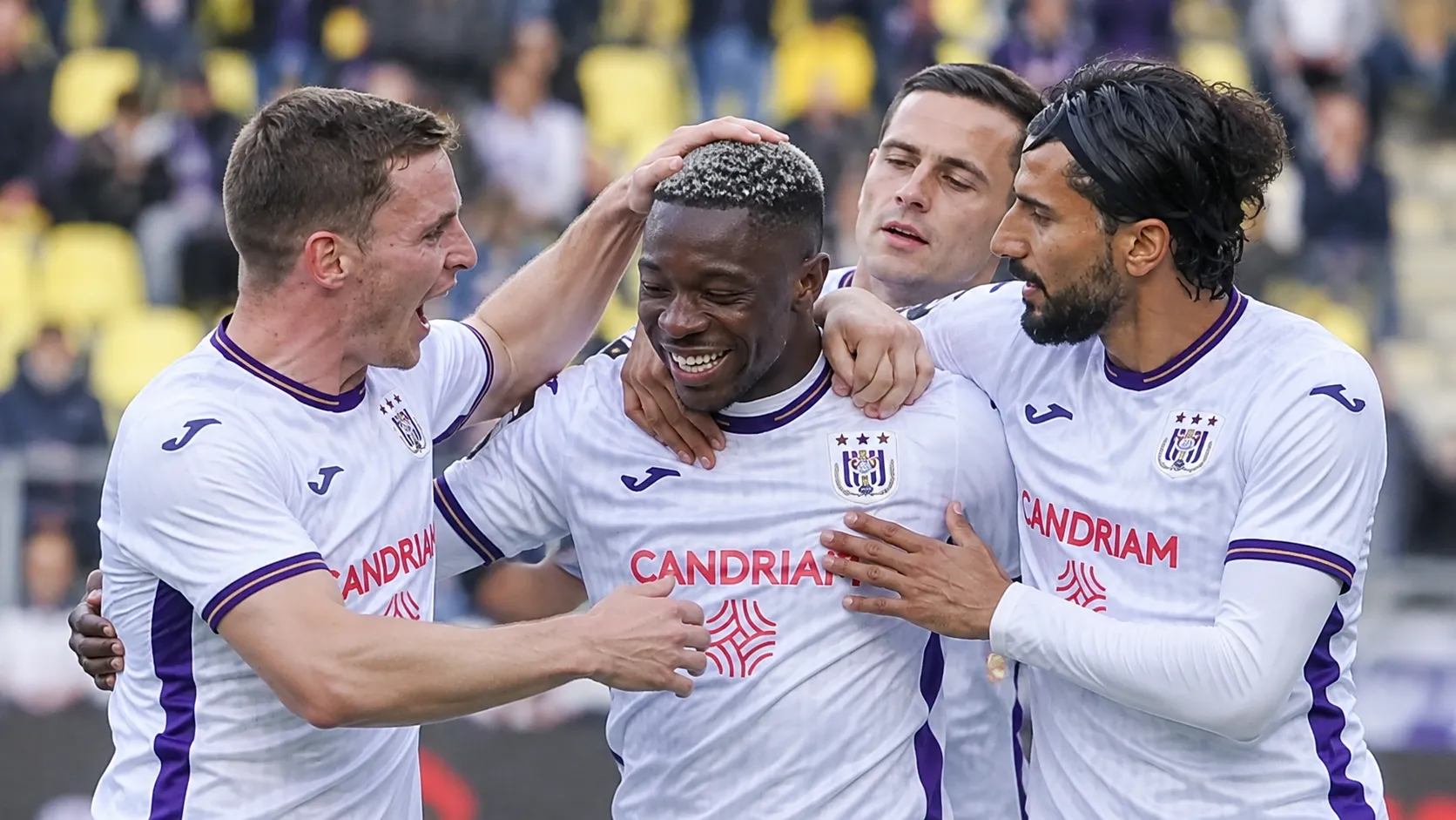 Een groep Anderlecht-spelers viert samen een doelpunt in witte shirts met paarse accenten.