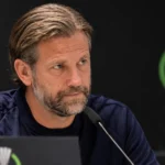 Een coach in donker trainingsshirt tijdens een persconferentie voor een donkere Conference League-achtergrond.