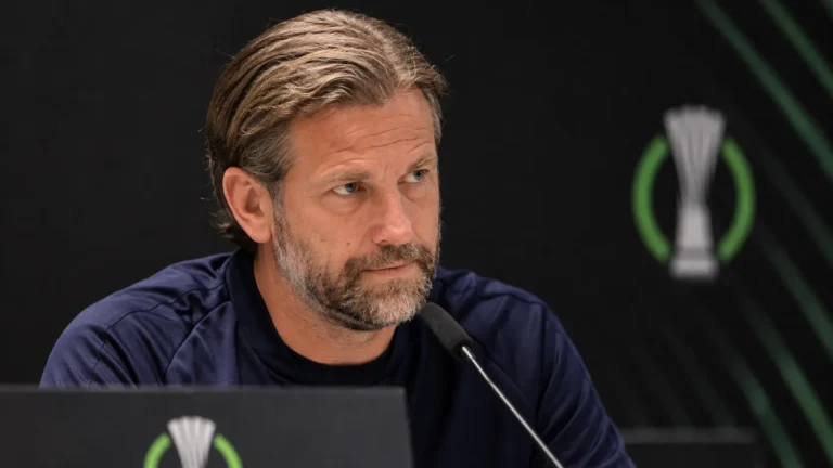 Een coach in donker trainingsshirt tijdens een persconferentie voor een donkere Conference League-achtergrond.